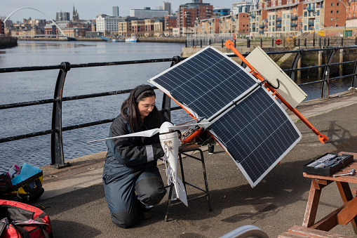 Female enginner working on a sustainable wind turbine fixed to a solar panel that can cahrage electric bikes. Millenuim bridge featuring in the background
