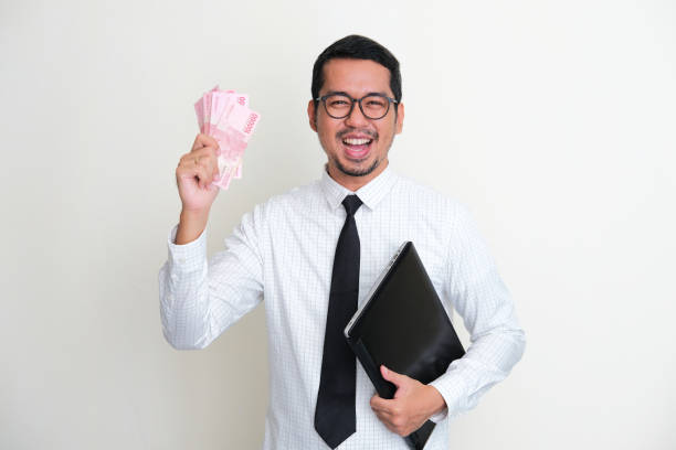 asiatischer geschäftsmann, der einen laptop und papiergeld in der hand hält und einen glücklichen ausdruck zeigt - indonesian currency stock-fotos und bilder