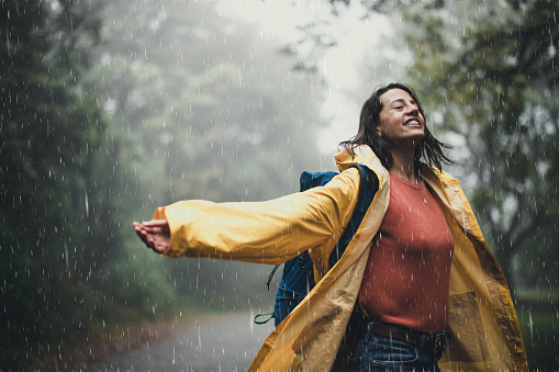 Young happy woman in yellow raincoat having fun during rainy day in the misty forest. Copy space.