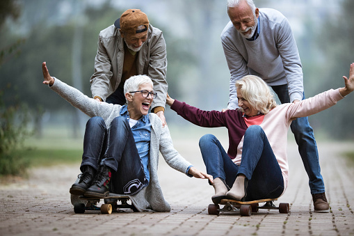 Happy mature women having fun while being pushed on skateboards by their husbands in the park.