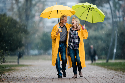 Happy senior couple in raincoats talking under umbrellas on a rainy day.