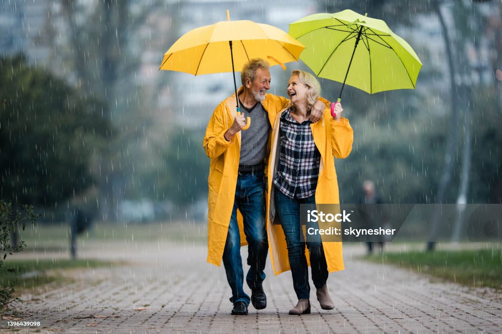 Happy senior couple in raincoats talking under umbrellas on a rainy day. Happy mature couple in yellow raincoats communicating under umbrellas on a rainy day. Raincoat Stock Photo