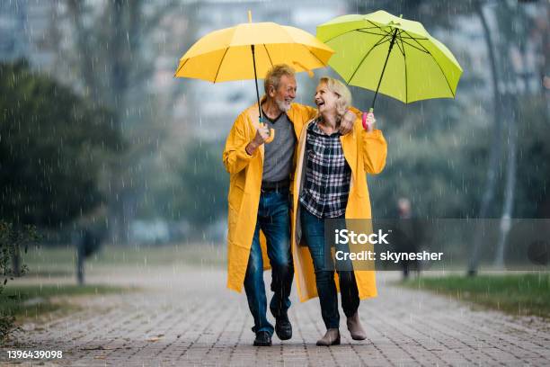 Happy Senior Couple In Raincoats Talking Under Umbrellas On A Rainy Day Stock Photo - Download Image Now