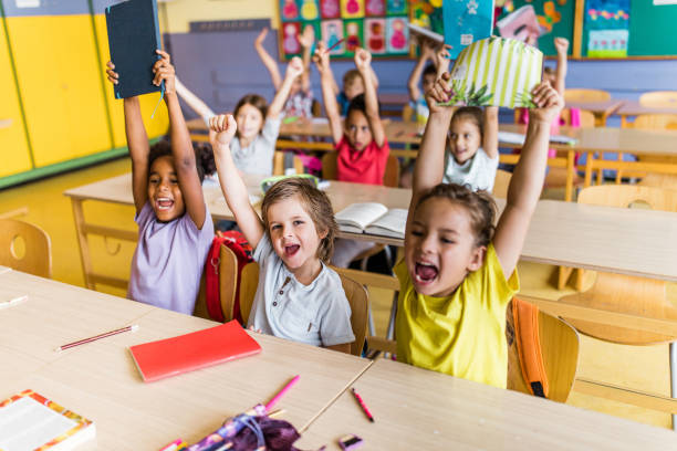 Large group of cheerful students celebrating the end of school year with raised arms in the classroom. Focus is on boy looking at camera.