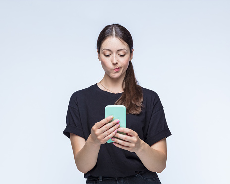 Angry young woman making face while using smart phone against white background.