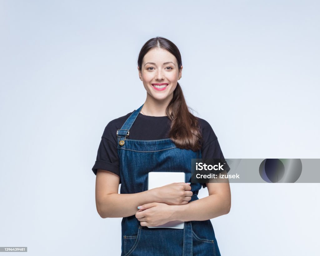 Happy woman standing with digital tablet Portrait of happy young woman wearing bib overalls standing with digital tablet against white background Waiter Stock Photo