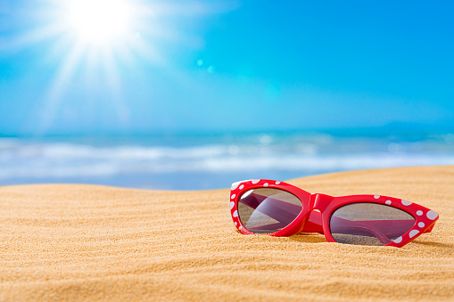 Red female sunglasses on golden tropical sandy beach. The composition is at the right of an horizontal frame leaving useful copy space for text and/or logo at the left. High resolution 42Mp outdoors digital capture taken with SONY A7rII and Zeiss Batis 40mm F2.0 CF lens
