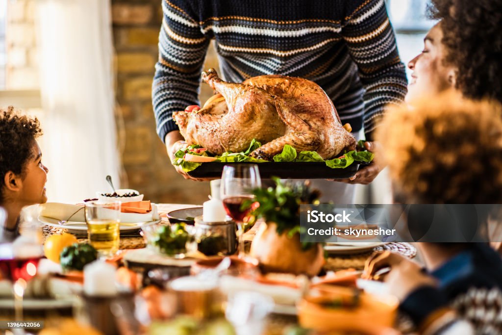 It is time for Thanksgiving turkey you all! Unrecognizable black father carrying Thanksgiving turkey while serving it for his family in dining room. Thanksgiving - Holiday Stock Photo