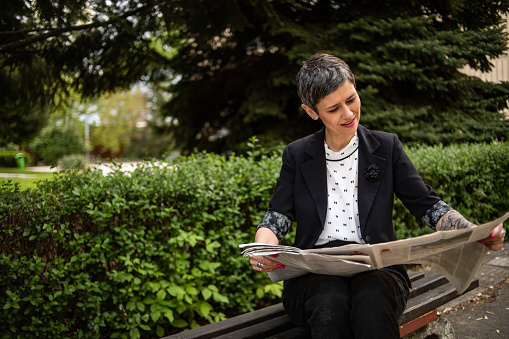 A mature woman is sitting on a park bench reading a newspaper