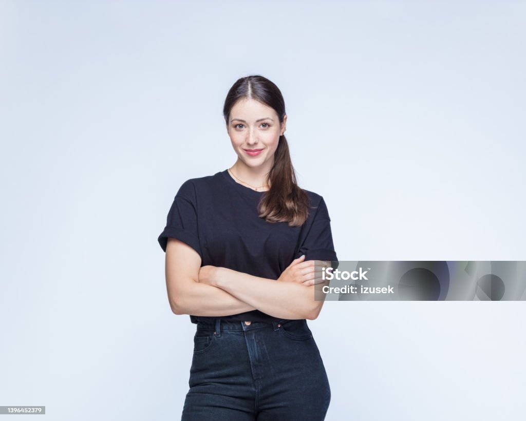 Woman with arms crossed against white background Portrait of smiling young woman wearing black clothes standing with arms crossed against white background. T-Shirt Stock Photo