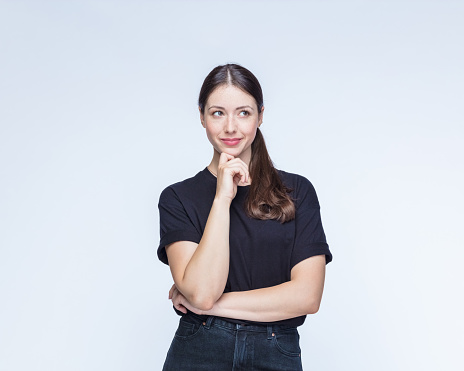 Young thoughtful woman wearing apron with hand on chin standing against white background.