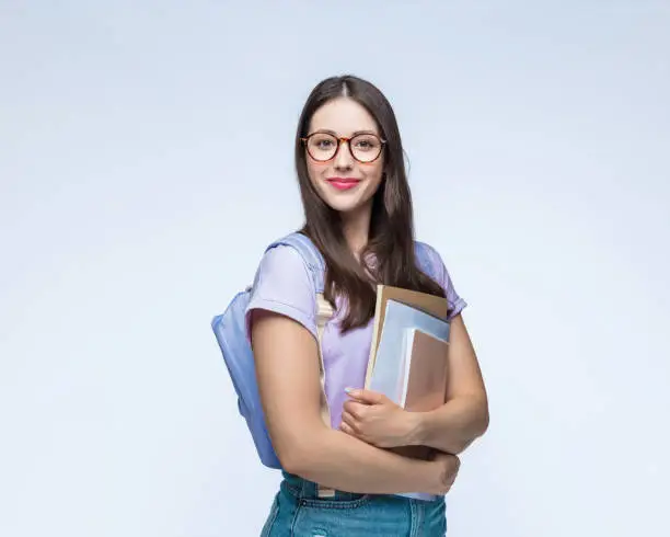 Portrait of confident young woman with books and backpack standing against white background.
