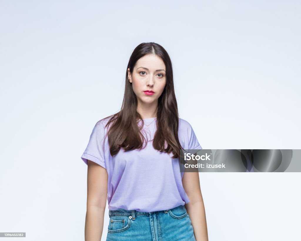 Woman in t-shirt with blank expression Portrait of young woman wearing lilac t-shirt with blank expression standing against white background. Portrait Stock Photo