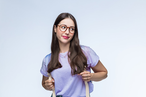Young woman with eyeglasses wearing lilac t-shirt carrying backpack against white background.