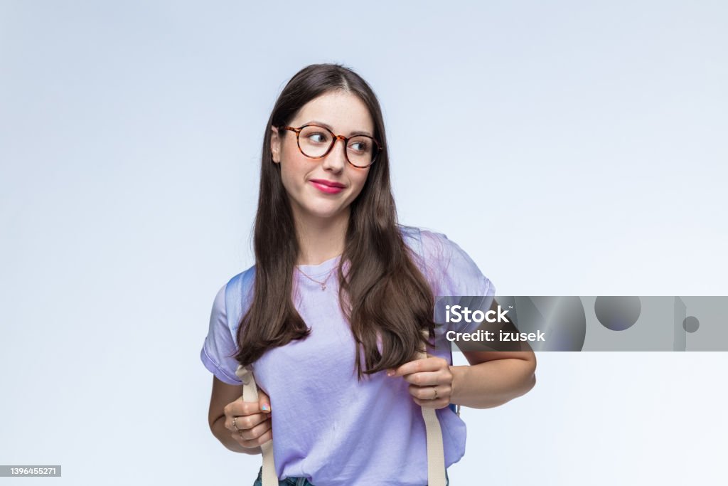 Woman looking away carrying backpack Young woman with eyeglasses wearing lilac t-shirt carrying backpack against white background. Looking Away Stock Photo