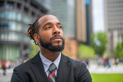 Headshot of a mid adult black male financial advisor looking away. He is with a serious face, wearing a black suit, a light color shirt and a tie. Defocused city buildings in the background.