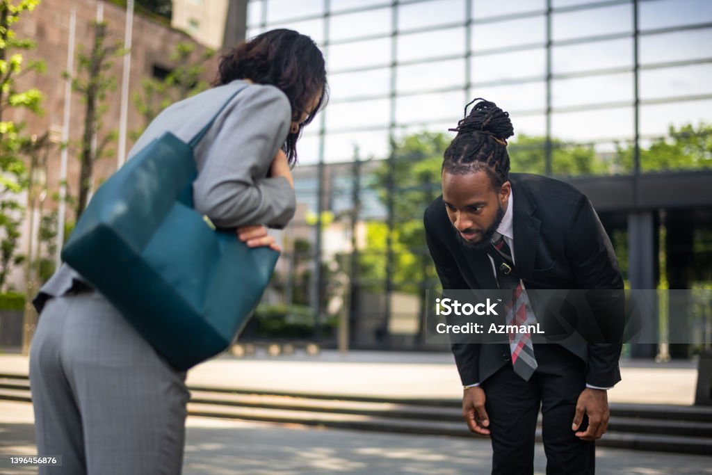 Black Male Lawyer And Japanese Female Client Bowing In Front Of A Building Japanese female businesswoman and black male lawyer bowing outdoors in front of a building. They are wearing formal business clothing. Defocused business building in the background. Bowing Stock Photo