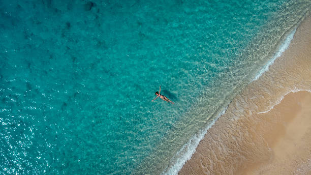 Aerial view of beautiful happy woman in swimsuit laying in the shallow sea water, enjoying sandy beach and soft turquoise ocean wave. Tropical sea in summer season on Egremni beach on Lefkada island. Aerial view of beautiful happy woman in swimsuit laying in the shallow sea water, enjoying sandy beach and soft turquoise ocean wave. Tropical sea in summer season on Egremni beach on Lefkada island. egremni beach lefkada island greece stock pictures, royalty-free photos & images