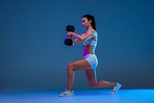 Portrait of yong muscular woman doing squats with heavy dumbbells, training isolated over blue studio background in neon light. Concept of sport, fitness, healthy lifestyle, strength, youth, ad