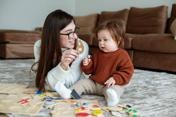 母と小さな娘は部屋の敷物の上で教育ゲームをします。茶色のセーターを着た幼児の赤ちゃん。 - 生後6ヶ月から11ヶ月 ストックフォトと画像