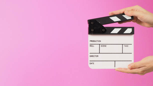 istock Two hands are holding a small white clapper board or movie slate on pink background. Side view 1396475630
