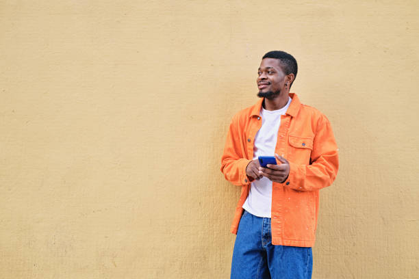 istock A young man holding his phone standing against an orange light wall looking away 1396476770