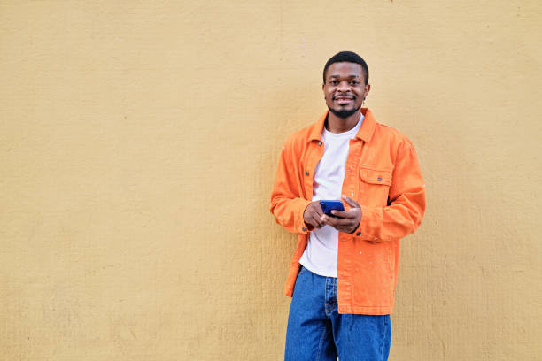 istock Standing young man holding his phone against an orange light wall looking at camera 1396476895