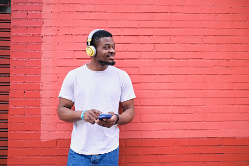 Front view of a young African American man in white shirt smiling holding his cell phone with a wireless headphones looking away against a red brick wall.