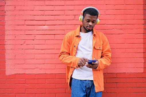 Front view of a young African American man in orange jacket listening to music with his wireless headphones and his phone against a red brick wall outdoors.
