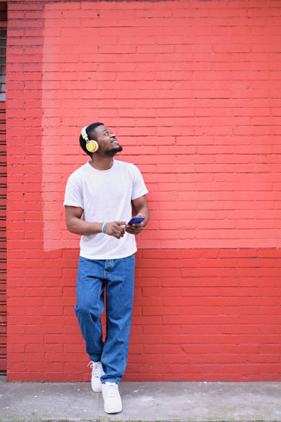 Vertical image of a young African American man in casual clothes leaning against a red brick wall using his cell phone with headphones looking up to copy space outdoors.