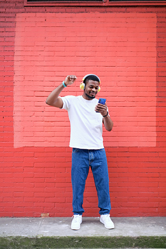 Vertical image of an enthusiastic young African American man listening to music on his cell phone and wireless headphones with one arm raised in the air against a red brick wall.