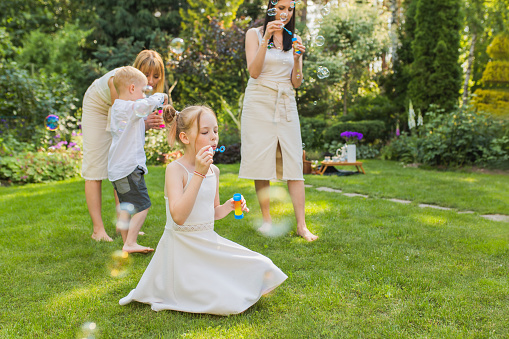 Blow and catch soap bubbles. Girl in white dress at backyard. Parents and children. Outdoor recreation. Childhood. Leisure activity.  Summer picnic vacation. Rest. Brothers and sisters. House rental