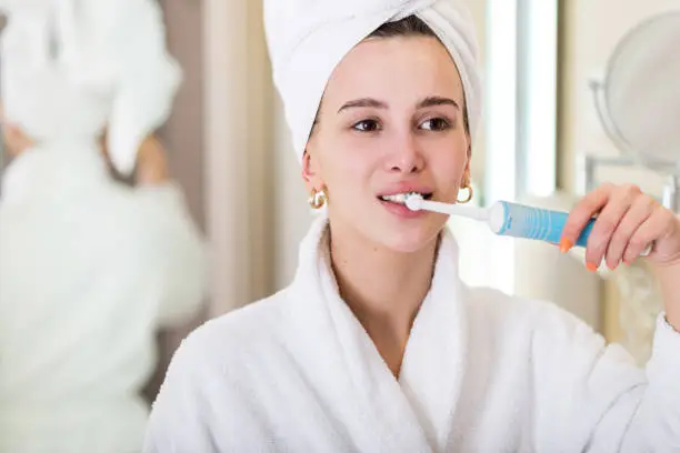 Attractive young woman brushing teeth at mirror wearing bathrobe