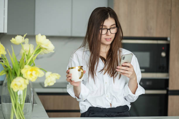 istock Beautiful woman is using smartphone in kitchen while drinking cup of freshly brewed coffee. 1396488167