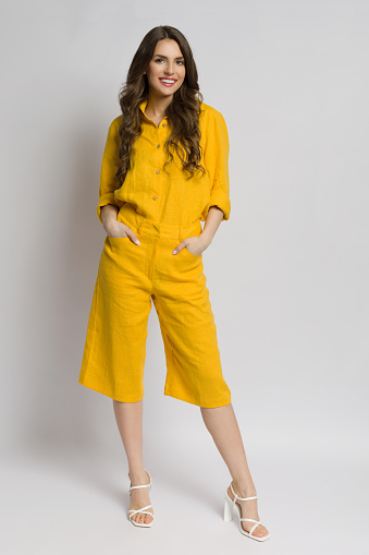 Beautiful young woman in yellow linen shirt, shorts and high heels poses with hands in pockets. Full length studio shot against white background.