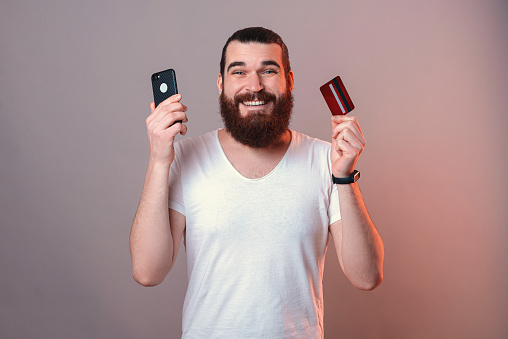 Wide smiling man is holding a credit card and phone near his face. Studio shot over grey background with red light.