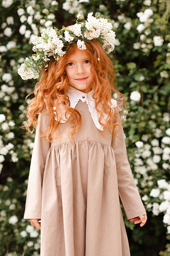 Cute little kid girl 3-4 year old with long curly red hair wear floral wreath and stylish rustic dress over nature background in garden outdoor. Springtime. Smiling baby with flowers. Childhood.