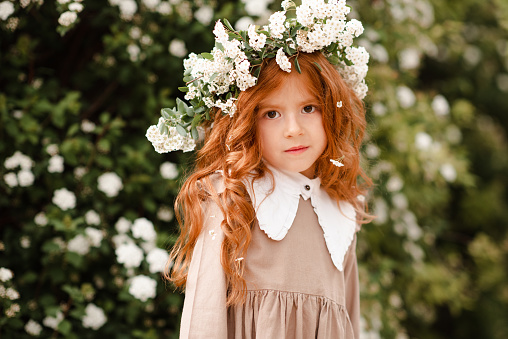 Cute little kid girl 3-4 year old with long curly red hair wear floral wreath and stylish rustic dress over nature background in garden outdoor. Springtime. Smiling baby with flowers. Childhood.