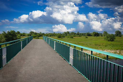 View of the bridge with metal railings. There are bushes behind the bridge, the sky with clouds