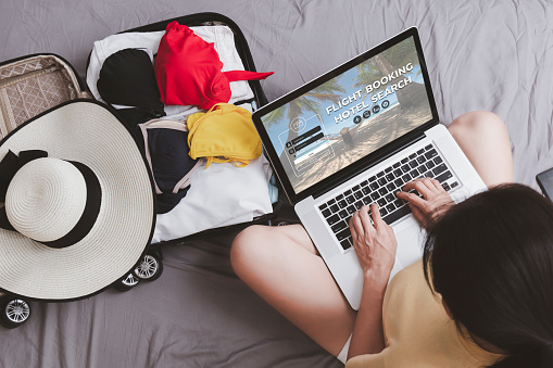 Top view Asian happy woman plan for trip by laptop booking hotel on the bed at home. Women with travel bag use notebook computer internet booking hotel. Holiday summer vacation concept