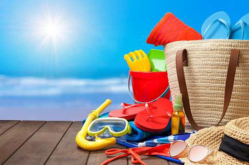 Beach bag with accessories and defocused sunny beach at background. The composition includes flip-flops, sand bucket, scuba mask, sunglasses, suntan lotion, sun hat, beach towel and parasol