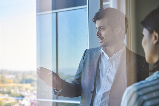 Side view shot of young adult businessman looking out the window while speaking with a woman inside the office