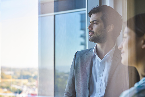 Side view portrait of two young adult business people looking out the office window with cityscape in the background