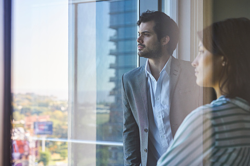 Side view portrait of two pensive young businesspeople standing next to the office window looking at the cityscape in the background