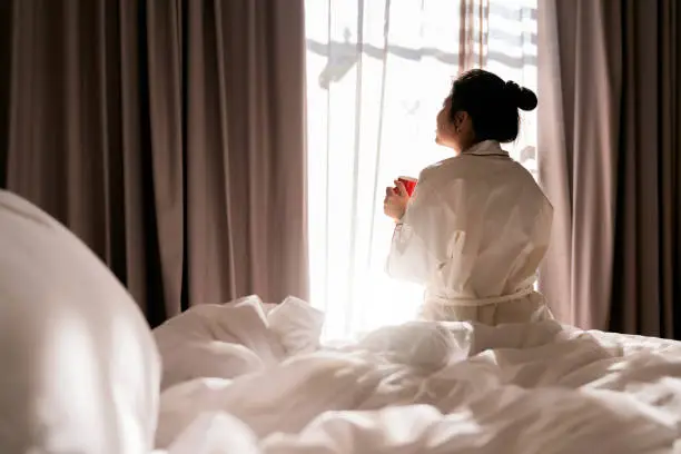Photo of Cheerful tender asain adult female in white casual cloth sitting with cup of coffee on bed and having happiness cheerful smiling enjoy freshness while enjoying morning in bedroom at home or hotel