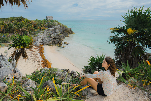 Young Caucasian woman  sitting and looking at  view of Tulum Mayan ruins at seaside