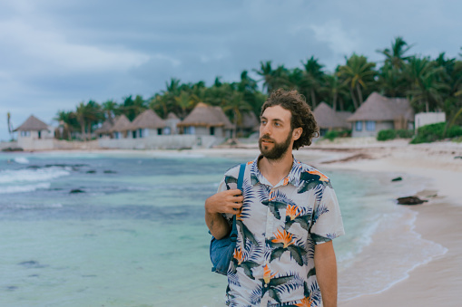 Young Caucasian man in Hawaiian shirt with backpack walking on the beach in Tulum