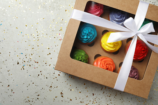 Box with delicious colorful cupcakes and confetti on light grey table, flat lay. Space for text