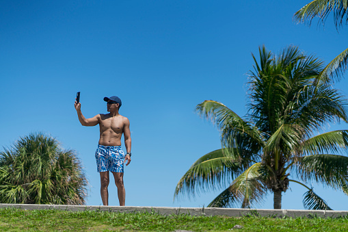Dark-skinned Latino man in his 30s wearing a sun hat and swimsuit shirtless on vacation at the beaches of Miami enjoying time alone while taking a selfie with his cell phone