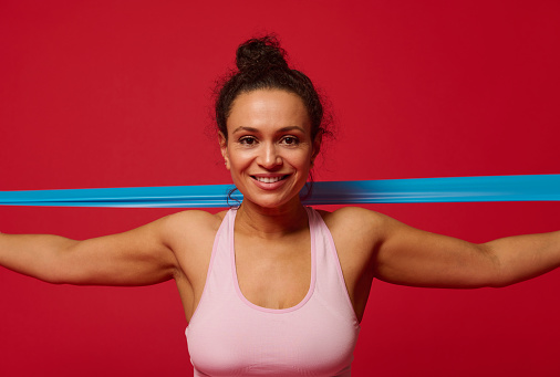 Close-up of a charming determined middle-aged sportswoman exercising with elastic fitness band, stretching her arms against red background with copy space for advertising text